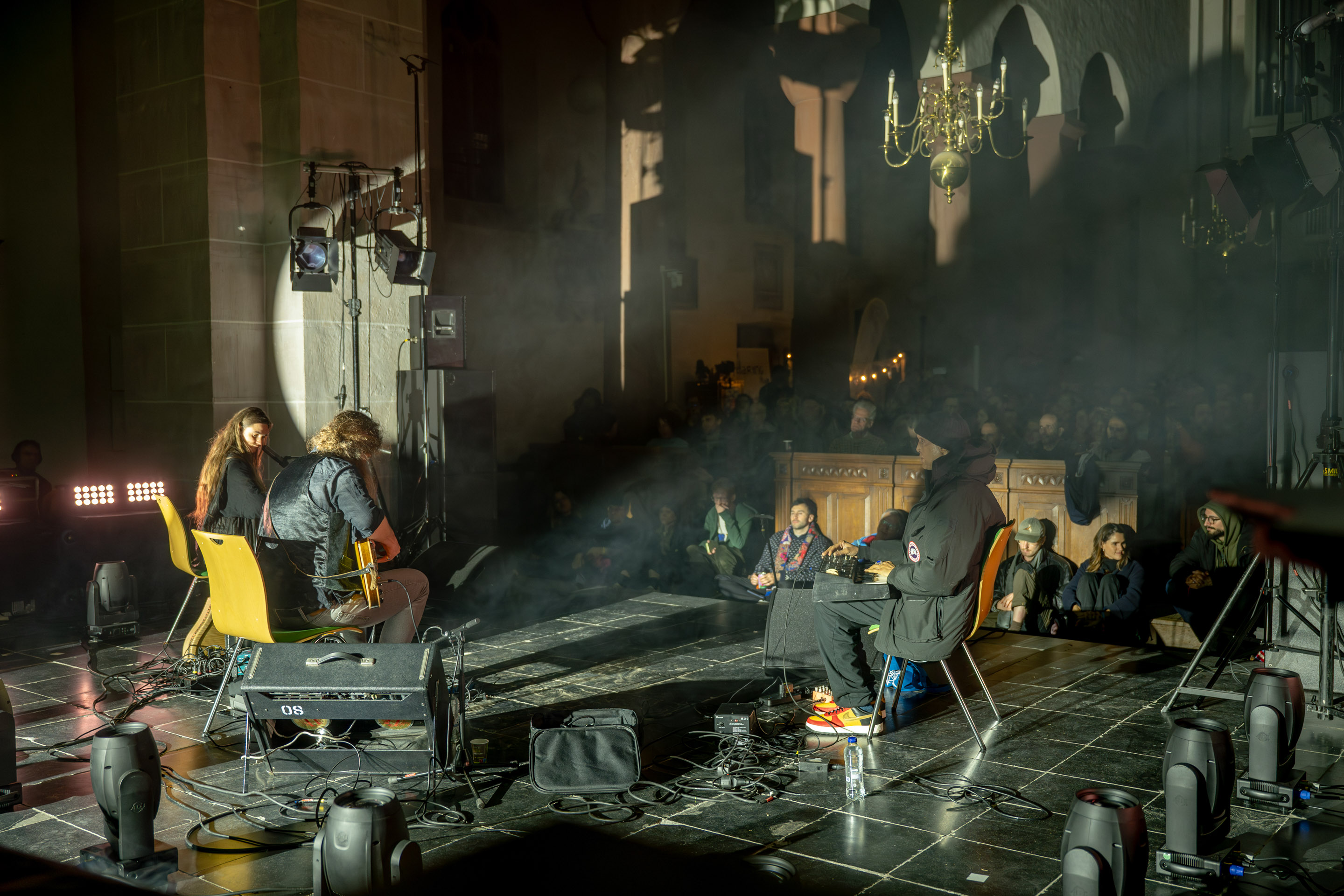 Three musicians are seated in a church while performing music. This photo is taken from behind the musicians and with audience seated in front of them.